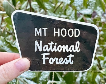 May include: A black and white sticker with a white border. The sticker has a wood grain texture and says "MT. HOOD National Forest" in white text.