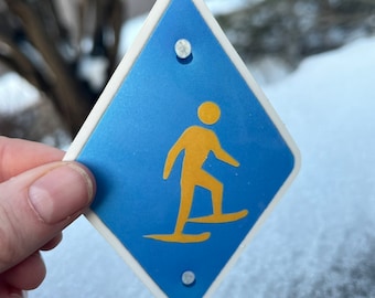 May include: A blue diamond-shaped sign with a white border and two white dots. The sign has a yellow silhouette of a person snowshoeing.