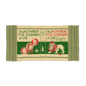 Peut inclure: Une serviette de plage rectangulaire avec la couverture de "The Wonderful Wizard of Oz". Le design comprend du texte vert et rouge et des illustrations de personnages comme l'Homme de fer-blanc et le Lion. La serviette a des bordures rayées vertes.