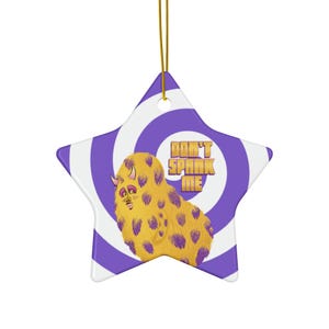 Puede incluir: Adorno en forma de estrella con un fondo en espiral morado y blanco. Presenta un monstruo amarillo con manchas moradas y el texto "DON'T SPANK ME" en dorado. Incluye un cordón dorado para colgar.