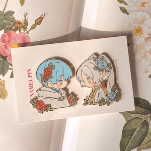 May include: Two enamel pins featuring stylised anime-style characters with floral accents on a white backing card. The left pin shows a character with blue hair and red roses, while the right pin depicts a character with white hair and blue roses.