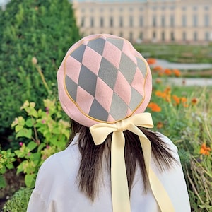 Könnte beinhalten: Eine rosa und graue karierte Baskenmütze mit goldener Einfassung und einem passenden gelben Band, das hinten zu einer Schleife gebunden ist.