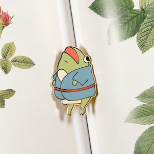 Könnte beinhalten: Ein skurriler Emaille-Pin mit einer Cartoon-Froschfigur, die einen blauen Bademantel und einen roten Hut trägt. Der Frosch hat einen grünen Körper, einen weißen Bauch und gelbe Beine. Der Pin befindet sich vor einem weißen Hintergrund mit Blumen- und Blätterillustrationen.
