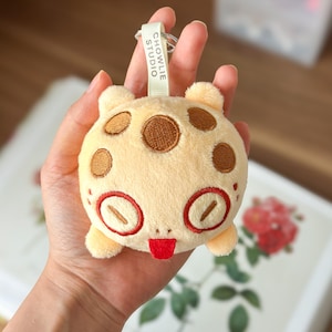 Pode incluir: Um porta-chaves de peluche redondo de cor creme com manchas circulares castanhas, olhos grandes com contorno vermelho e uma pequena língua vermelha de fora. Uma etiqueta de tecido branco no anel metálico exibe o texto CHOWLIE STUDIO.