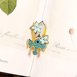Könnte beinhalten: Eine goldfarbene Emaille-Anstecknadel mit einem stilisierten Schwert mit blauer Klinge und gelbem Griff. Das Schwert ist mit weißen Blumen und einem grünen Blatt verziert. Die Anstecknadel befindet sich auf einer Buchseite mit französischem Text.