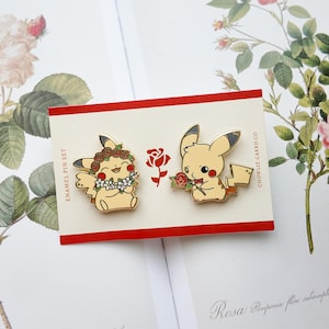 Könnte beinhalten: Zwei goldene Emaille-Pins mit einem Cartoon-Pikachu. Ein Pikachu trägt eine Blumenkrone und das andere hält eine rote Rose. Die Pins befinden sich auf einer roten und weißen Karte mit dem Text "Enamel Pin Set" und "Chowlie Card Co."
