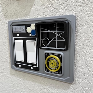 May include: A gray wall-mounted control panel with two white toggle switches, a small screen with a black and white design, and a yellow circular button with a black and white design.