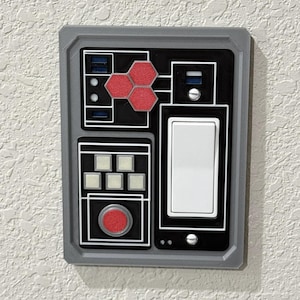 May include: A gray wall switch plate with a black and white design. The plate has a red button, a light switch, and a panel with a red hexagonal pattern.