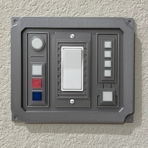 Puede incluir: Una placa de interruptor de pared gris con un interruptor de luz blanco y varios botones más pequeños. Los botones son rojos, azules y blancos.