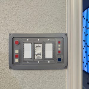 Puede incluir: Una placa de interruptor de pared gris con temática de Star Wars. Tiene tres interruptores de palanca, dos de los cuales son blancos y uno es un atenuador. La placa también tiene un botón rojo y un botón azul.