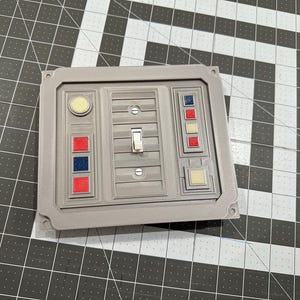 Puede incluir: Una cubierta de interruptor de luz gris, rectangular, con un diseño retro. Cuenta con un interruptor basculante central, múltiples botones de colores y un botón circular. La cubierta probablemente está impresa en 3D y tiene una estética de ciencia ficción o industrial.