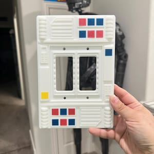 May include: White plastic light switch cover with a futuristic design. It has blue and red squares and a yellow rectangle.