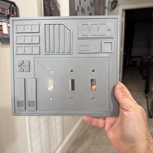Puede incluir: Una tapa de interruptor de luz gris impresa en 3D con un diseño de Star Wars. La tapa tiene un panel con botones y un panel con tres interruptores.