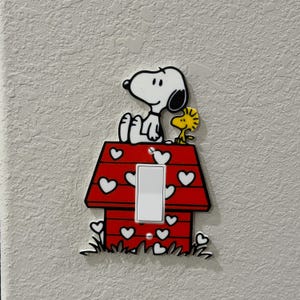 Peut inclure: Un cache-interrupteur décoratif avec Snoopy et Woodstock. Snoopy est assis sur une niche rouge ornée de cœurs blancs. Woodstock se tient à côté de Snoopy. L'interrupteur est blanc et rectangulaire.
