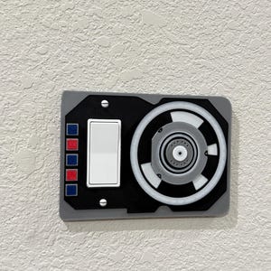May include: A gray and black wall switch with a round, white, and gray speaker. The switch has a white toggle and four colored buttons.
