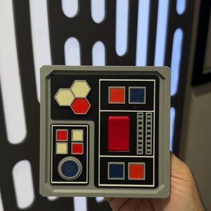 May include: A gray, square panel with a black interior featuring various colored geometric shapes and buttons. The panel has red, blue, and yellow accents. The background is a black and white pattern.