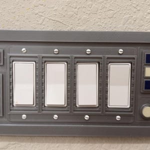 May include: A gray wall-mounted panel with five white toggle switches, a red button, and three black buttons. The panel has a silver trim and a circular button in the lower right corner.