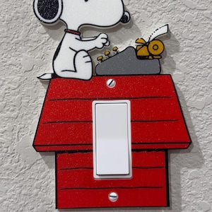 Puede incluir: Una placa de interruptor roja con forma de caseta de perro con un perro blanco con gorra naranja sentado encima, escribiendo en una máquina de escribir gris. El diseño incluye un interruptor blanco central y dos tornillos plateados de montaje.