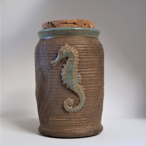 以下が含まれることがあります： コルクの蓋と青いタツノオトシゴのデザインが施された茶色の陶器の壺。壺は表面がざらざらしていて、上部に薄い青色の釉薬がかけられている。