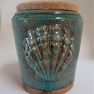 May include: A teal ceramic jar with a cork lid and a raised seashell design. The jar has a speckled glaze and a brown rim.