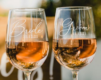 May include: Two clear wine glasses with the words "Bride" and "Groom" etched on them. The glasses are filled with a light pink beverage and the date "11.12.21" is etched below each word.