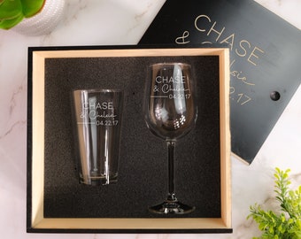 Puede incluir: Una caja de regalo de madera con tapa negra. La tapa tiene el texto "& CHASE" grabado. La caja contiene dos vasos, una copa de vino y una copa de pinta. Ambos vasos tienen el texto "CHASE & Chelsea 04.22.17" grabado.