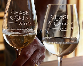 Puede incluir: Dos copas de vino transparentes llenas de un líquido claro. Cada copa está grabada con los nombres "Chase & Chelsie" y la fecha "02.22.17". Las copas son sostenidas por una persona, con un fondo borroso.