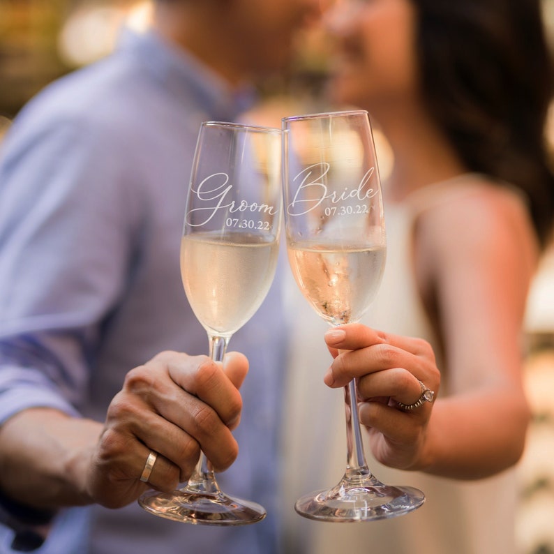 Etched stemmed champagne glasses. There is a set of two in the image; one glass says Groom in cursive and a date below. The other glass says Bride in cursive and a date below.