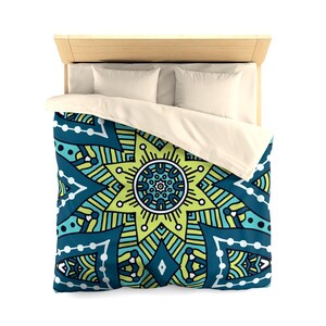 Può includere: Un letto con struttura in legno, due cuscini bianchi e un copripiumino con un motivo mandala geometrico turchese, verde e giallo. Il copripiumino è ripiegato sul letto, mostrando il design intricato.