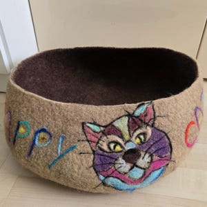 Puede incluir: Una cama para gatos de fieltro beige con interior marrón. El exterior presenta una cara de gato colorida y la palabra "Happy" bordada en azul y amarillo. La cama tiene forma de cuenco redondo y abierto.