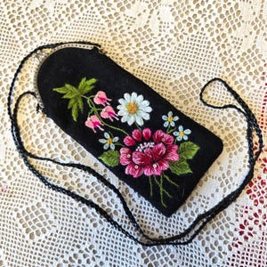 Op de afbeelding: Zwarte vilten crossbodytas met een bloemenborduurontwerp. De borduursels hebben roze, witte en blauwe bloemen met groene bladeren. De tas heeft een zwarte gevlochten riem en een zilveren sluiting.