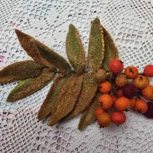 May include: A handcrafted felted botanical arrangement featuring brown and green leaves and clusters of orange and red berries. The arrangement is likely a decorative item, possibly for fall or autumn-themed decor.