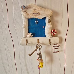 May include: A wall-mounted key holder shaped like a blue door with a white driftwood frame. It features a Welcome sign, a small bell, a lifebuoy, and seashells. Three metal hooks hold a brass key and a small yellow mouse figurine keychain.