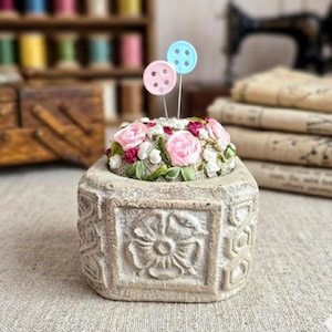 May include: A stone-effect pin cushion with a floral design. The cushion is adorned with pink and red roses, white flowers, and green ribbon. Two button-shaped pins, one pink and one blue, are inserted into the cushion.