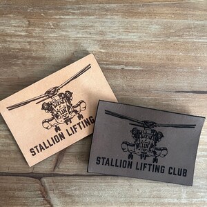 May include: Two rectangular leather patches, one tan and one gray, with a black helicopter graphic and text. The tan patch reads "STALLION LIFTING", and the gray patch reads "STALLION LIFTING CLUB".