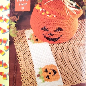 May include: A crocheted orange pumpkin with a black smiling face, holding candy corn. The pumpkin is sitting on a white and brown crocheted mat with a smaller pumpkin design. The text "11 Trick or Treat" is in the top left corner of the image.
