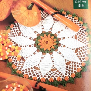 May include: White crochet doily with orange and green accents. The doily is on a wooden surface with orange pumpkins and candy corn.