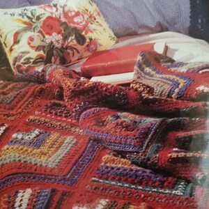 May include: A red, orange, green and blue crocheted throw blanket with a floral cushion and a red book on top of it.