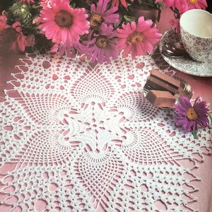 Puede incluir: Doily de crochet blanco con un delicado patrón floral, colocado sobre una superficie rosa con una taza y un platillo y cuadrados de chocolate.
