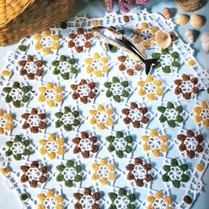 May include: A white crocheted doily with a repeating pattern of brown, green, and yellow flowers. The doily is on a light blue surface.