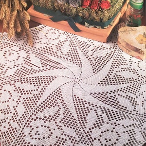 May include: A white crocheted doily with a circular design. The doily is laid out on a wooden table with a bouquet of dried flowers and a small decorative heart-shaped tile in the background.