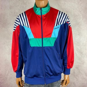 May include: A vintage Adidas track jacket in blue, red, and teal. The jacket features white stripes on the sleeves and a green collar. The Adidas logo is visible on the chest. The jacket is displayed on a mannequin.