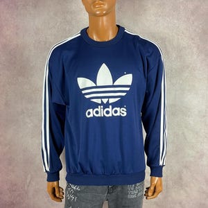 May include: Navy blue Adidas sweatshirt with white stripes on the sleeves and a large white Adidas logo on the front. The sweatshirt has a crew neck and a ribbed hem. A black baseball cap with a yellow emblem is worn on the mannequin's head.