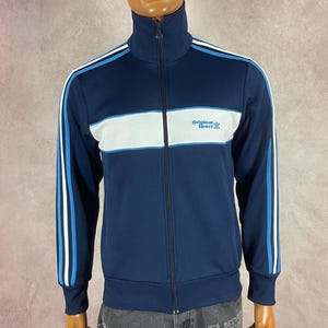 May include: Navy blue track jacket with white and light blue stripes. The jacket has a high collar and a full-length zipper. The text "Originals Sport" is printed on the chest. A black cap with a yellow emblem is worn on the mannequin's head.