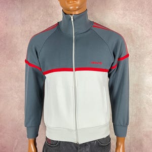 May include: A gray, white, and red track jacket with a full zipper. The jacket has red stripes on the shoulders and a red logo on the chest. A red baseball cap is on the mannequin's head.