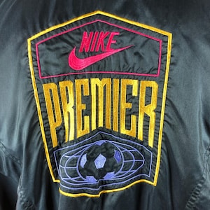 Puede incluir: Chaqueta negra con el logo "NIKE PREMIER" bordado en dorado, rosa y rojo. El logo incluye un gr&aacute;fico de bal&oacute;n de f&uacute;tbol en azul y morado. La chaqueta est&aacute; hecha de un material brillante.