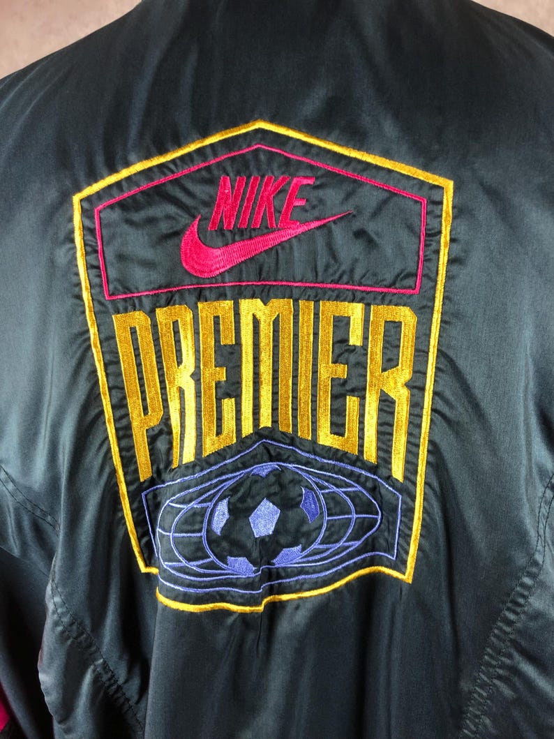 Puede incluir: Chaqueta negra con el logo "NIKE PREMIER" bordado en dorado, rosa y rojo. El logo incluye un gr&aacute;fico de bal&oacute;n de f&uacute;tbol en azul y morado. La chaqueta est&aacute; hecha de un material brillante.