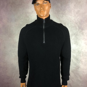 May include: A black ribbed knit sweater with a half-zip closure, displayed on a mannequin. The sweater has a high collar and long sleeves. The mannequin is also wearing a navy blue baseball cap with a yellow logo.