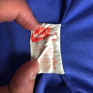 Puede incluir: Primer plano de una prenda azul con una etiqueta blanca. La etiqueta presenta el logotipo de Nike en rojo y el texto "FUPAS". El texto de la etiqueta indica la composici&oacute;n del tejido, incluyendo nailon y poli&eacute;ster.