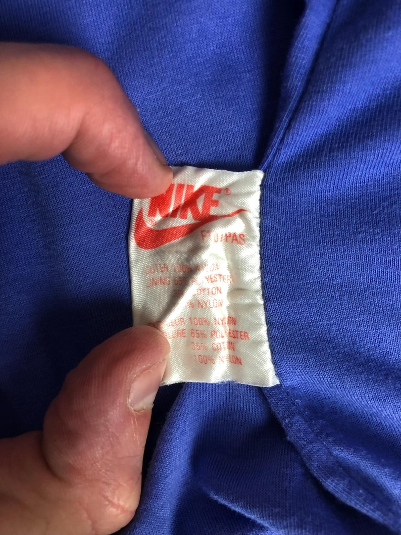 Puede incluir: Primer plano de una prenda azul con una etiqueta blanca. La etiqueta presenta el logotipo de Nike en rojo y el texto "FUPAS". El texto de la etiqueta indica la composici&oacute;n del tejido, incluyendo nailon y poli&eacute;ster.